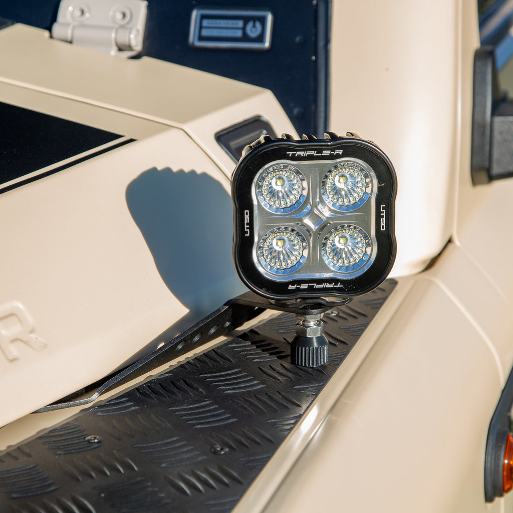 A powder-coated black aluminum Ditch Light Bracket and LED Triple-R Brand Off-road LED light mounted on the edge of the hood with no drilling needed, showing a Spacer to reduce vibration and bouncing of the light. 
LED ditch light attached to a vehicle's hood. 